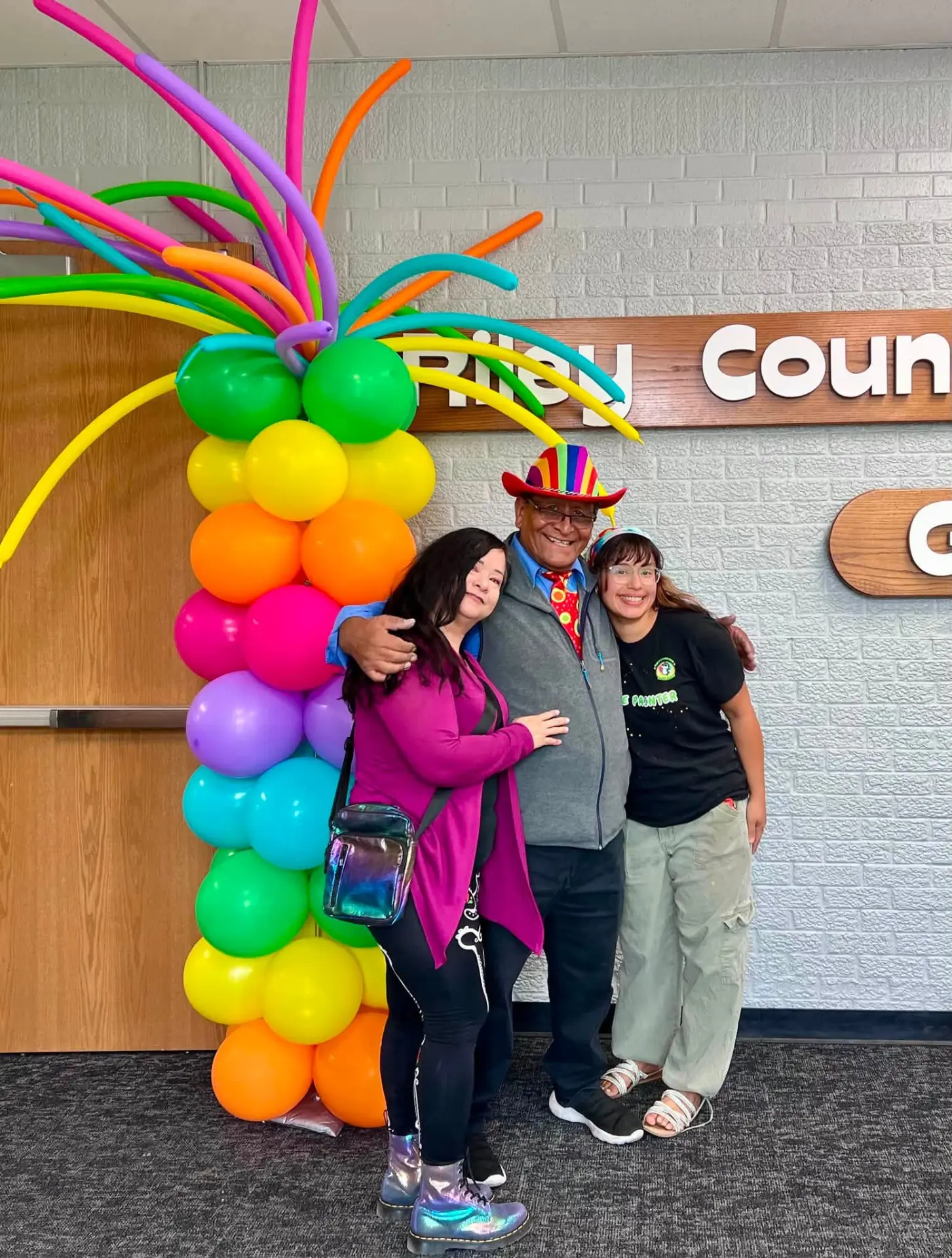 Carnival Corner entertainers smiling beside a rainbow balloon column at a community event