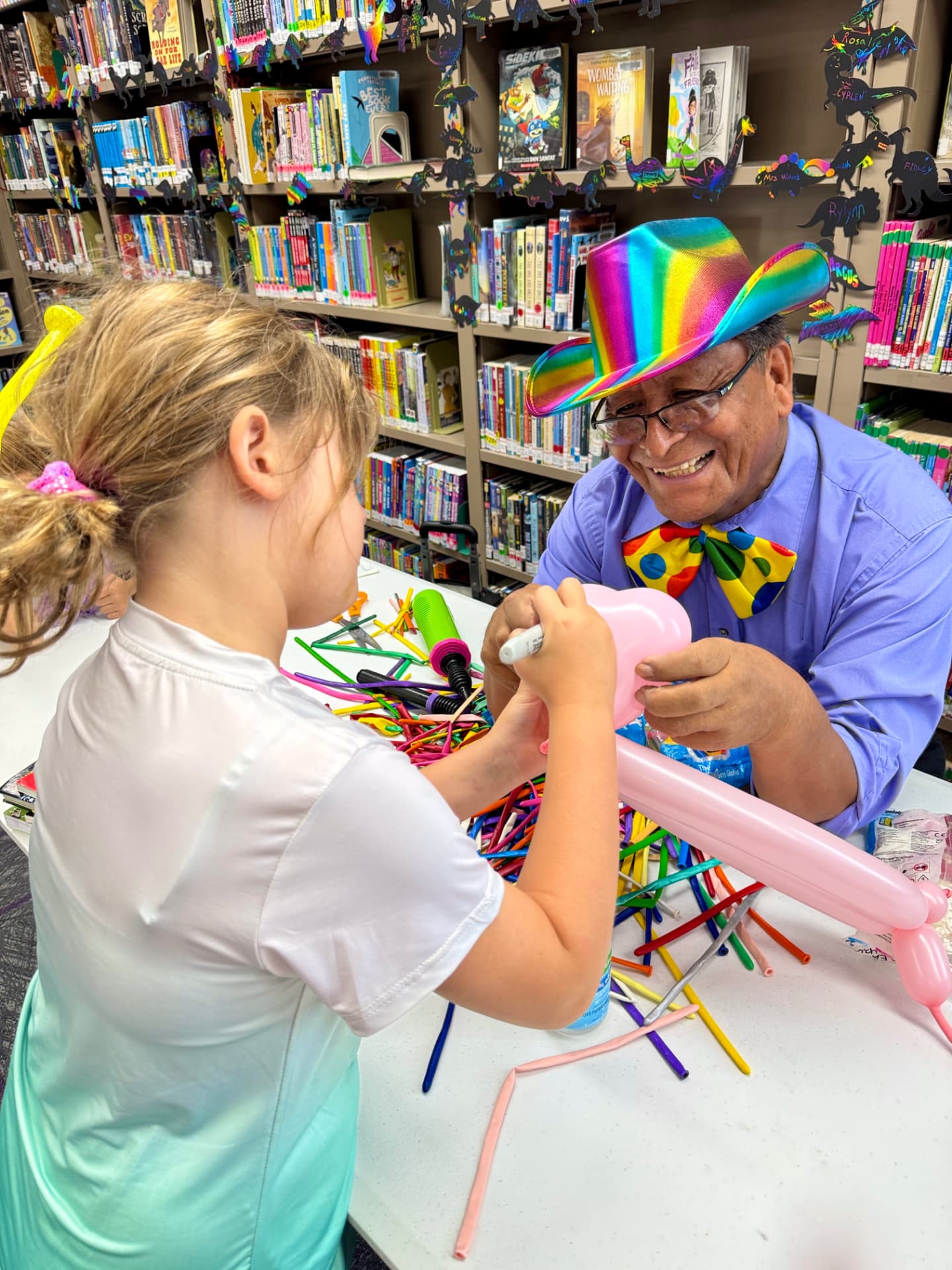 Balloon artist in a rainbow hat helping a young girl decorate a balloon at a library workshop